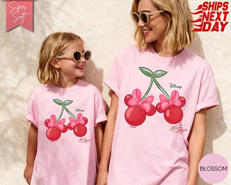 May include: Two light pink t-shirts featuring a cherry design with Minnie Mouse bows and the Disney logo. The shirts are worn by a woman and a child, both wearing sunglasses. The shirts are labeled "Super Soft" and "Blossom".