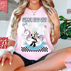 May include: A light pink t-shirt featuring a graphic print of Minnie Mouse sitting on a white unicorn pool float. The design includes palm trees, sunglasses, a popsicle, and the text SUMMER VIBES ONLY above a black and white checkered pattern.