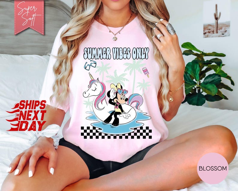 May include: A light pink t-shirt featuring a graphic print of Minnie Mouse sitting on a white unicorn pool float. The design includes palm trees, sunglasses, a popsicle, and the text SUMMER VIBES ONLY above a black and white checkered pattern.