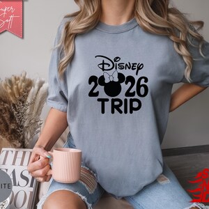 May include: A light blue t-shirt with the text "Disney 2026 TRIP" in black. The design includes a Minnie Mouse silhouette with a bow. The shirt is a soft, comfortable material. The words "Super Soft" are on a pink banner.