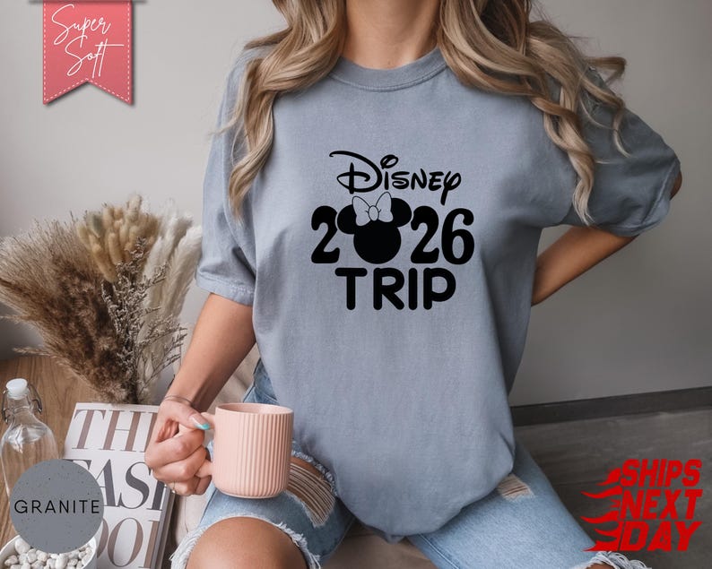 May include: A light blue t-shirt with the text "Disney 2026 TRIP" in black. The design includes a Minnie Mouse silhouette with a bow. The shirt is a soft, comfortable material. The words "Super Soft" are on a pink banner.