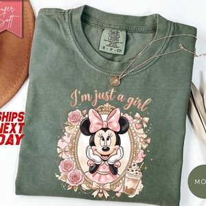 Może przedstawiać: Mchowo-zielony t-shirt z grafiką Myszki Minnie w ozdobnej złotej ramce, otoczonej r&oacute;żowymi r&oacute;żami, kokardkami i kubkiem kawy. Tekst nad ramką brzmi I'm just a girl. Koszulka posiada okrągły dekolt oraz kr&oacute;tkie rękawy i jest wykonana z bawełny.