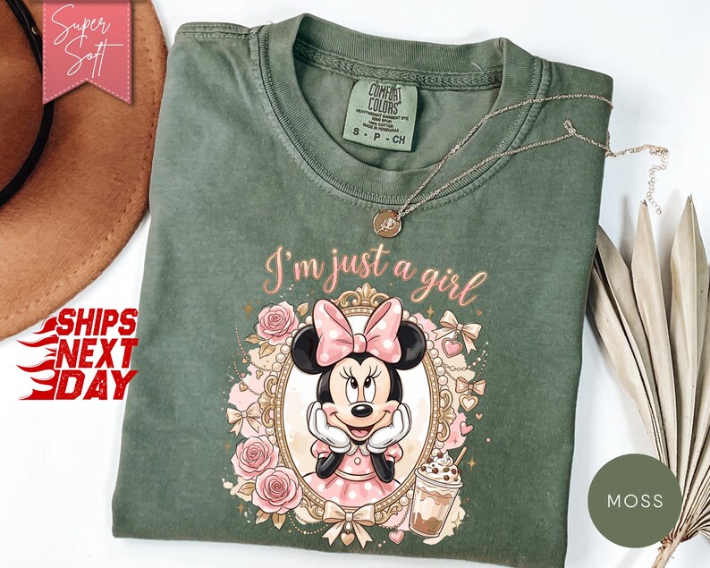 Może przedstawiać: Mchowo-zielony t-shirt z grafiką Myszki Minnie w ozdobnej złotej ramce, otoczonej r&oacute;żowymi r&oacute;żami, kokardkami i kubkiem kawy. Tekst nad ramką brzmi I'm just a girl. Koszulka posiada okrągły dekolt oraz kr&oacute;tkie rękawy i jest wykonana z bawełny.