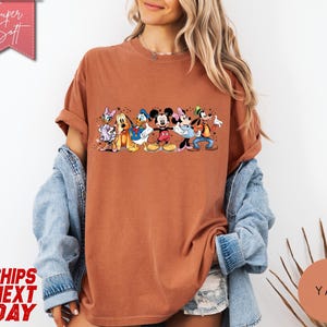 Puede incluir: Una camiseta de color &oacute;xido con un gr&aacute;fico de personajes de Disney, incluyendo Mickey y Minnie Mouse, Donald Duck y Goofy. La camiseta tiene las mangas enrolladas y el texto "Super Soft" es visible en una pancarta rosa.