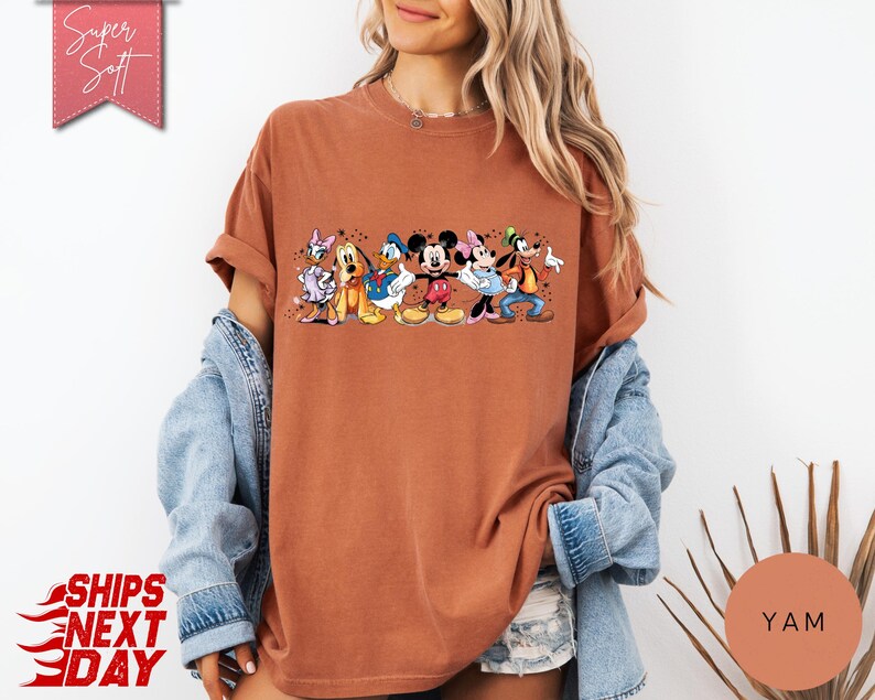 Puede incluir: Una camiseta de color &oacute;xido con un gr&aacute;fico de personajes de Disney, incluyendo Mickey y Minnie Mouse, Donald Duck y Goofy. La camiseta tiene las mangas enrolladas y el texto "Super Soft" es visible en una pancarta rosa.