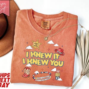 May include: A terracotta-coloured crew neck t-shirt featuring a graphic print with the text I KNEW IT I KNEW YOU, a sheriff star badge labelled INFINITY AND BEYOND, a red cowboy hat, cowboy boots, a beaded bracelet, and stacked alphabet blocks.