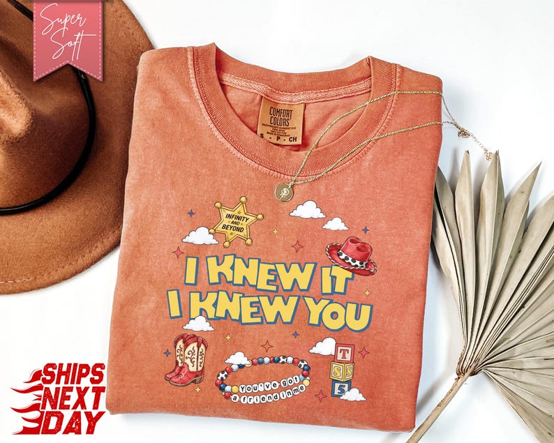 May include: A terracotta-coloured crew neck t-shirt featuring a graphic print with the text I KNEW IT I KNEW YOU, a sheriff star badge labelled INFINITY AND BEYOND, a red cowboy hat, cowboy boots, a beaded bracelet, and stacked alphabet blocks.