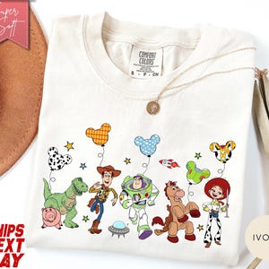 May include: An ivory t-shirt featuring a graphic print of Toy Story characters including Woody, Buzz Lightyear, Rex, Hamm, Bullseye, and Jessie. The characters are holding Mickey Mouse-shaped balloons. The shirt has a Comfort Colors brand label.