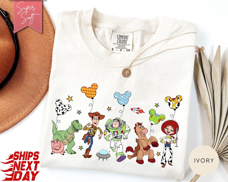 May include: An ivory t-shirt featuring a graphic print of Toy Story characters including Woody, Buzz Lightyear, Rex, Hamm, Bullseye, and Jessie. The characters are holding Mickey Mouse-shaped balloons. The shirt has a Comfort Colors brand label.