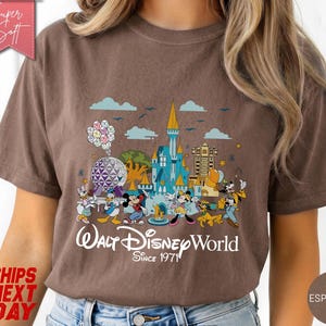 K&ouml;nnte beinhalten: Braunes T-Shirt mit einer farbenfrohen Grafik von Walt Disney World-Charakteren und -Wahrzeichen. Das Design zeigt Micky Maus, Donald Duck und ein Schloss mit dem Text "Walt Disney World Since 1971".