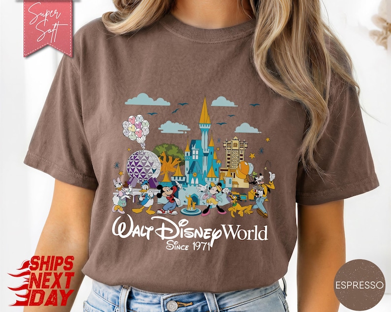 K&ouml;nnte beinhalten: Braunes T-Shirt mit einer farbenfrohen Grafik von Walt Disney World-Charakteren und -Wahrzeichen. Das Design zeigt Micky Maus, Donald Duck und ein Schloss mit dem Text "Walt Disney World Since 1971".