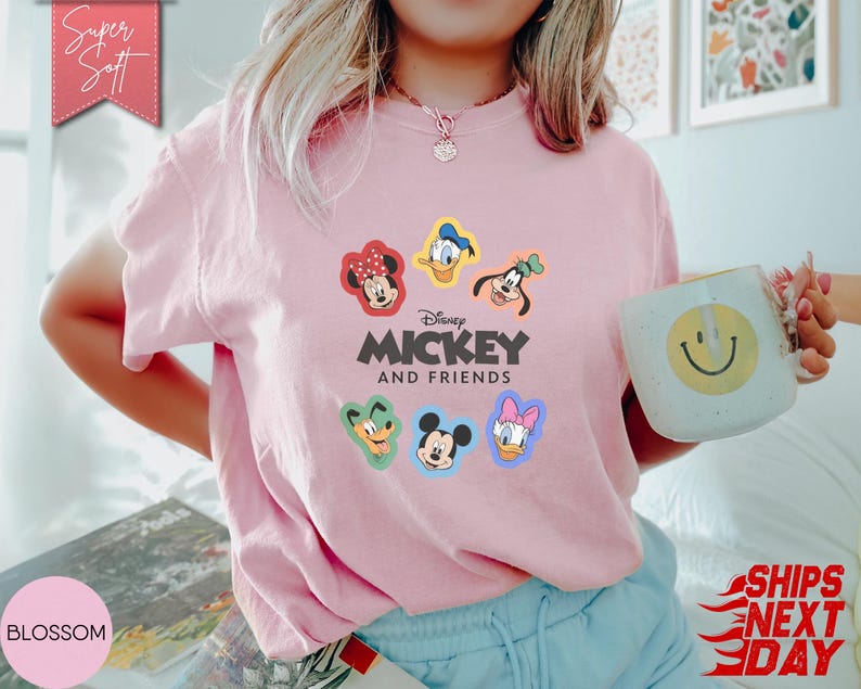 May include: A light pink t-shirt featuring the text "Disney Mickey and Friends" with colorful cartoon character graphics. The shirt has a crew neck and short sleeves. A mug with a smiley face is held in the hand.