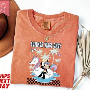 May include: A terracotta-colored t-shirt featuring a graphic of a cartoon mouse sitting on a unicorn pool float in water, surrounded by palm trees, sunglasses, and a popsicle. The text reads "SUMMER VIBES ONLY" above a black and white checkerboard pattern.