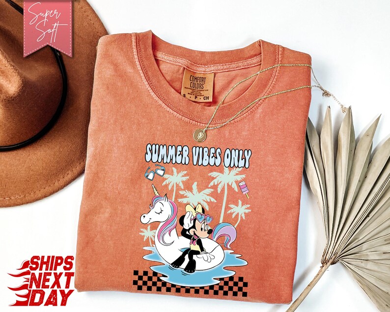 May include: A terracotta-colored t-shirt featuring a graphic of a cartoon mouse sitting on a unicorn pool float in water, surrounded by palm trees, sunglasses, and a popsicle. The text reads "SUMMER VIBES ONLY" above a black and white checkerboard pattern.