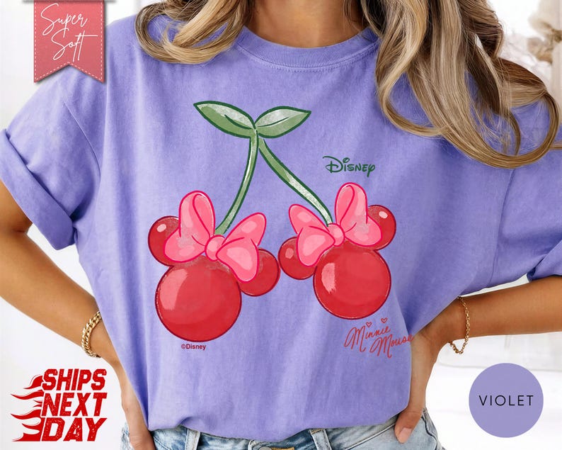 May include: Violet t-shirt featuring a Minnie Mouse-inspired cherry design. The cherries are red with pink bows, green leaves, and the Disney logo. The shirt has rolled-up sleeves and a relaxed fit.