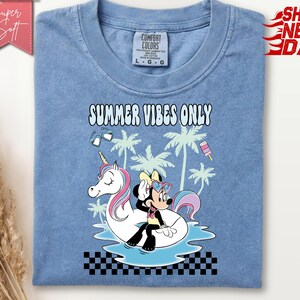 May include: A blue t-shirt featuring a graphic of Minnie Mouse sitting on a white unicorn pool float. The design includes palm trees, heart-shaped sunglasses, a popsicle, and the text "SUMMER VIBES ONLY" above a black and white checkered border.