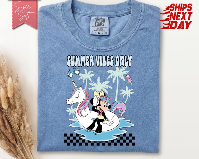 May include: A blue t-shirt featuring a graphic of Minnie Mouse sitting on a white unicorn pool float. The design includes palm trees, heart-shaped sunglasses, a popsicle, and the text "SUMMER VIBES ONLY" above a black and white checkered border.