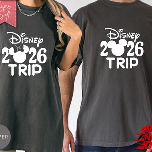 May include: Two dark gray t-shirts with the text "Disney 2026 TRIP" in white. One shirt features a Minnie Mouse bow. The shirts are made of a soft material and are suitable for a Disney vacation.