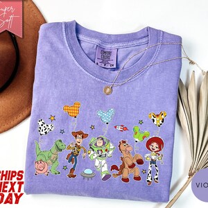 May include: A light purple crew neck t-shirt featuring a graphic print of Toy Story characters including Woody, Buzz Lightyear, Rex, Hamm, Bullseye, and Jessie holding Mickey Mouse-shaped balloons, with a Comfort Colors brand label at the neck.