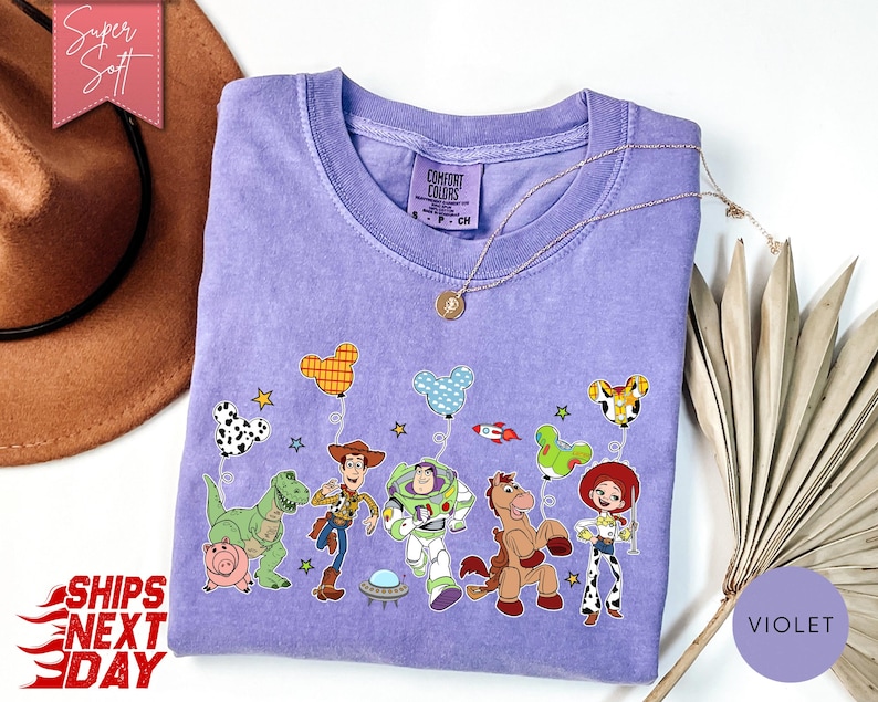 May include: A light purple crew neck t-shirt featuring a graphic print of Toy Story characters including Woody, Buzz Lightyear, Rex, Hamm, Bullseye, and Jessie holding Mickey Mouse-shaped balloons, with a Comfort Colors brand label at the neck.