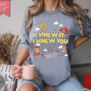 May include: A slate blue short-sleeve t-shirt featuring a graphic print with the text I KNEW IT I KNEW YOU, a yellow sheriff star, a red cowboy hat, red cowboy boots, a beaded bracelet, and three stacked alphabet blocks with the letters T, S, and S.