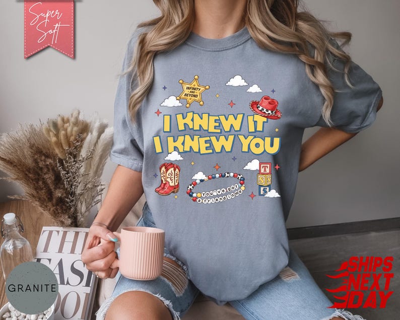 May include: A slate blue short-sleeve t-shirt featuring a graphic print with the text I KNEW IT I KNEW YOU, a yellow sheriff star, a red cowboy hat, red cowboy boots, a beaded bracelet, and three stacked alphabet blocks with the letters T, S, and S.