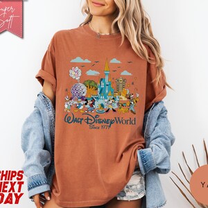K&ouml;nnte beinhalten: Rostfarbenes T-Shirt mit einer farbenfrohen Grafik des Walt Disney World Schlosses, Figuren und dem Text "Walt Disney World Since 1971." Das Shirt hat hochgekrempelte &Auml;rmel und eine lockere Passform.