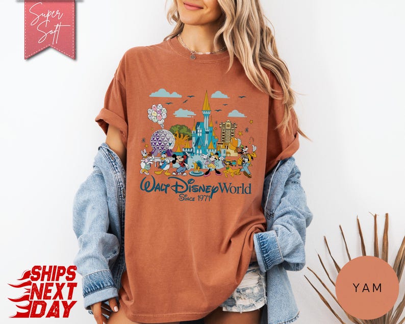 K&ouml;nnte beinhalten: Rostfarbenes T-Shirt mit einer farbenfrohen Grafik des Walt Disney World Schlosses, Figuren und dem Text "Walt Disney World Since 1971." Das Shirt hat hochgekrempelte &Auml;rmel und eine lockere Passform.