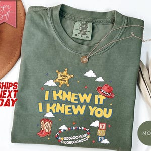 May include: A moss green short-sleeve t-shirt featuring a graphic print with the text I KNEW IT I KNEW YOU, a gold sheriff star badge with the text INFINITY AND BEYOND, a red cowboy hat, red cowboy boots, a beaded bracelet, and alphabet blocks.
