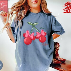 May include: Blue jean colored t-shirt featuring a cartoon cherry design with pink bows and green stems. The shirt has the Disney logo and the words "Minnie Mouse". The words "Super Soft" and "Ships Next Day" are also visible.