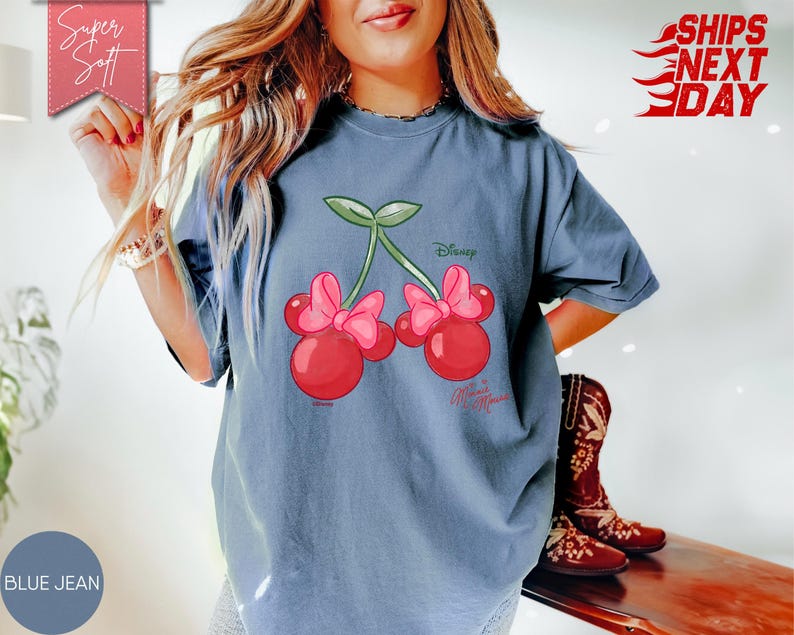 May include: Blue jean colored t-shirt featuring a cartoon cherry design with pink bows and green stems. The shirt has the Disney logo and the words "Minnie Mouse". The words "Super Soft" and "Ships Next Day" are also visible.