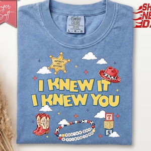 May include: A blue crew neck t-shirt featuring a graphic print with the text I KNEW IT I KNEW YOU. The design includes a yellow sheriff star badge with the text INFINITY AND BEYOND, a red cowboy hat, red cowboy boots, a beaded bracelet, and blocks.