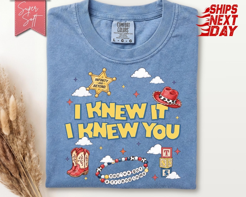 May include: A blue crew neck t-shirt featuring a graphic print with the text I KNEW IT I KNEW YOU. The design includes a yellow sheriff star badge with the text INFINITY AND BEYOND, a red cowboy hat, red cowboy boots, a beaded bracelet, and blocks.