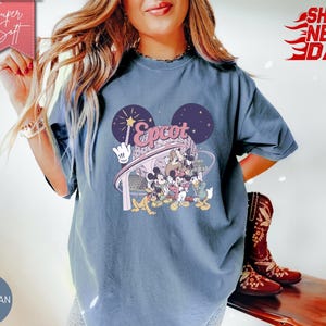 May include: Blue jean colored t-shirt featuring a graphic of Disney characters and the word "Epcot" in a stylized font. The design includes Mickey Mouse ears and a wand. The shirt is a relaxed fit.