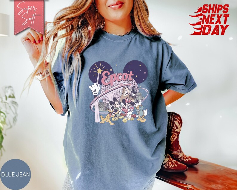 May include: Blue jean colored t-shirt featuring a graphic of Disney characters and the word "Epcot" in a stylized font. The design includes Mickey Mouse ears and a wand. The shirt is a relaxed fit.