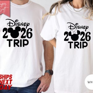May include: White t-shirts with a black Disney design. The design includes the word "Disney", the numbers "2026", a Mickey Mouse head, and the word "TRIP". One shirt has a bow on the Mickey Mouse head. The shirts are labeled "Super Soft".