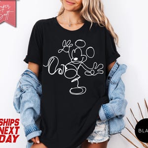 May include: Black t-shirt featuring a white line drawing of Mickey Mouse with the text "Love" and "Ours". The shirt has short sleeves and is worn with a denim jacket. The words "Super Soft" and "Ships Next Day" are also visible.