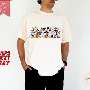 Puede incluir: Camiseta de color marfil con un gr&aacute;fico colorido de personajes de Disney, incluyendo Mickey Mouse, Donald Duck y Goofy. La camiseta tiene mangas cortas y cuello redondo. Las palabras "Super Soft" est&aacute;n en una pancarta rosa.