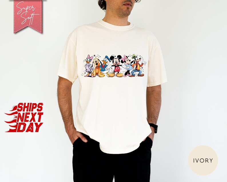 Puede incluir: Camiseta de color marfil con un gr&aacute;fico colorido de personajes de Disney, incluyendo Mickey Mouse, Donald Duck y Goofy. La camiseta tiene mangas cortas y cuello redondo. Las palabras "Super Soft" est&aacute;n en una pancarta rosa.