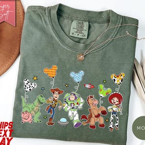 May include: A moss green t-shirt featuring a graphic print of Toy Story characters including Rex, Hamm, Woody, Buzz Lightyear, Bullseye, and Jessie. The characters are holding Mickey Mouse-shaped balloons, with small stars and a rocket ship.