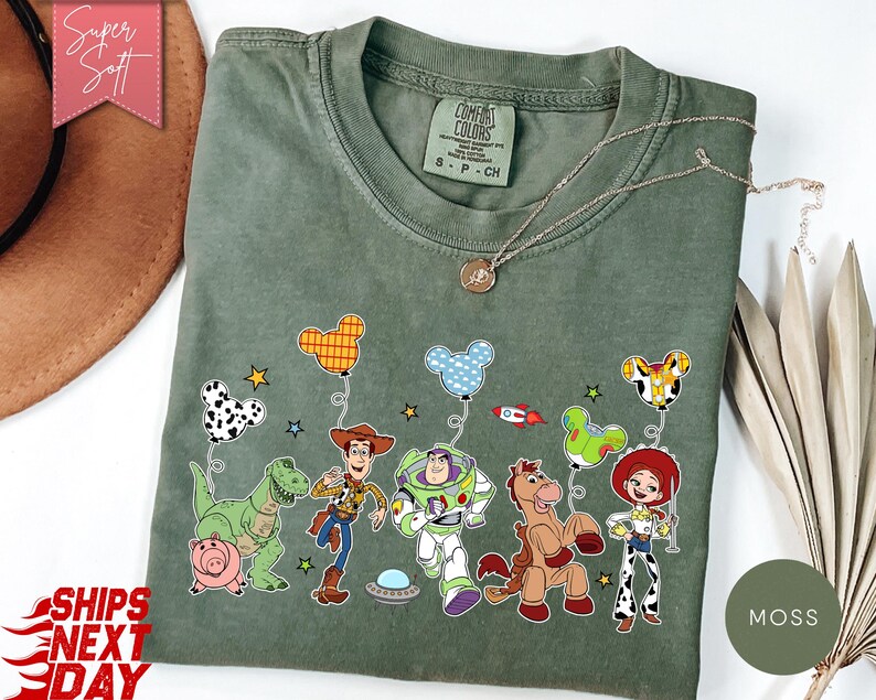 May include: A moss green t-shirt featuring a graphic print of Toy Story characters including Rex, Hamm, Woody, Buzz Lightyear, Bullseye, and Jessie. The characters are holding Mickey Mouse-shaped balloons, with small stars and a rocket ship.