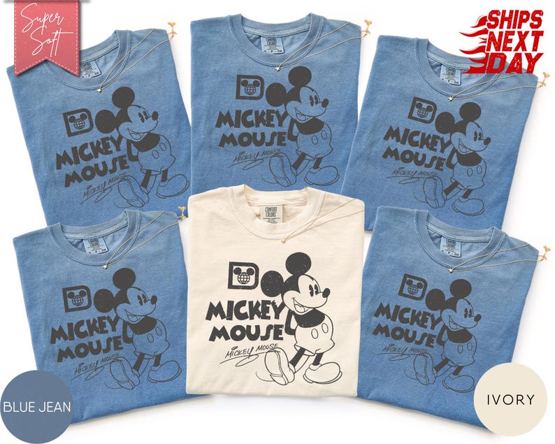 May include: A collection of blue jean and ivory-colored t-shirts featuring a classic Mickey Mouse graphic. Each shirt displays "Mickey Mouse" in a bold font, with a smiling Mickey Mouse illustration. The shirts are adorned with a delicate necklace.