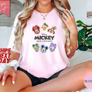May include: A light pink t-shirt featuring the text "Mickey and Friends" with cartoon character graphics. The shirt has a crew neck and short sleeves. The image also includes the text "Super Soft" and "Ships Next Day".