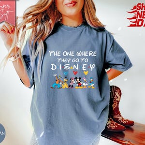 K&ouml;nnte beinhalten: Blaues Jeans-T-Shirt mit dem Text "The One Where They Go To Disney" und Disney-Figuren. Das Shirt hat einen Rundhalsausschnitt und kurze &Auml;rmel. Die Worte "Super Soft" stehen in einem rosa Banner.