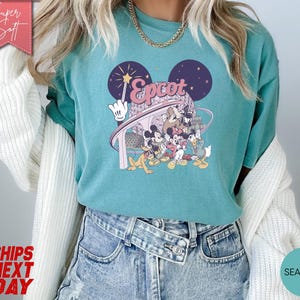 May include: Teal-colored t-shirt featuring a graphic of Disney characters, including Mickey Mouse, Donald Duck, and Goofy, with the word "Epcot" in pink. The design includes a wand and a stylized depiction of Spaceship Earth. The shirt is made of a soft material.