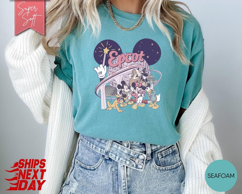 May include: Teal-colored t-shirt featuring a graphic of Disney characters, including Mickey Mouse, Donald Duck, and Goofy, with the word "Epcot" in pink. The design includes a wand and a stylized depiction of Spaceship Earth. The shirt is made of a soft material.