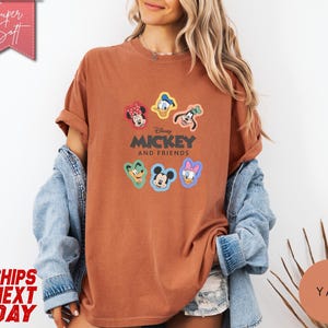 May include: A rust-colored t-shirt featuring the text "Mickey and Friends" with cartoon character graphics. The shirt has rolled-up sleeves and is worn with a denim jacket. The image also includes the text "Super Soft" and "Ships Next Day".