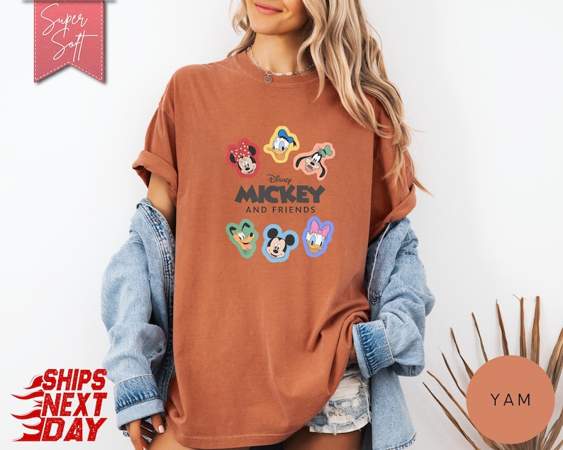 May include: A rust-colored t-shirt featuring the text "Mickey and Friends" with cartoon character graphics. The shirt has rolled-up sleeves and is worn with a denim jacket. The image also includes the text "Super Soft" and "Ships Next Day".