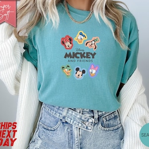 May include: Seafoam green t-shirt featuring the text "Disney Mickey and Friends" with cartoon character faces. The shirt has a crew neck and short sleeves. The shirt is made of a soft material.