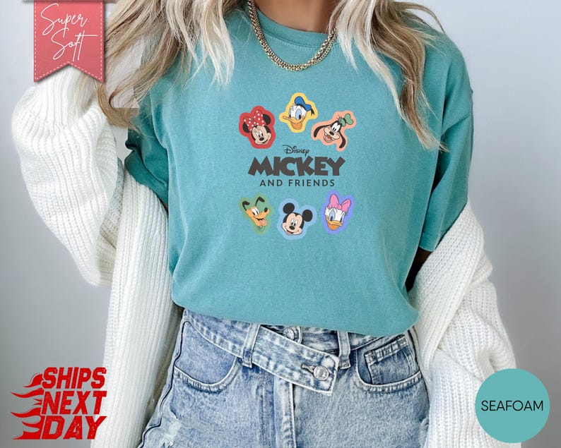 May include: Seafoam green t-shirt featuring the text "Disney Mickey and Friends" with cartoon character faces. The shirt has a crew neck and short sleeves. The shirt is made of a soft material.
